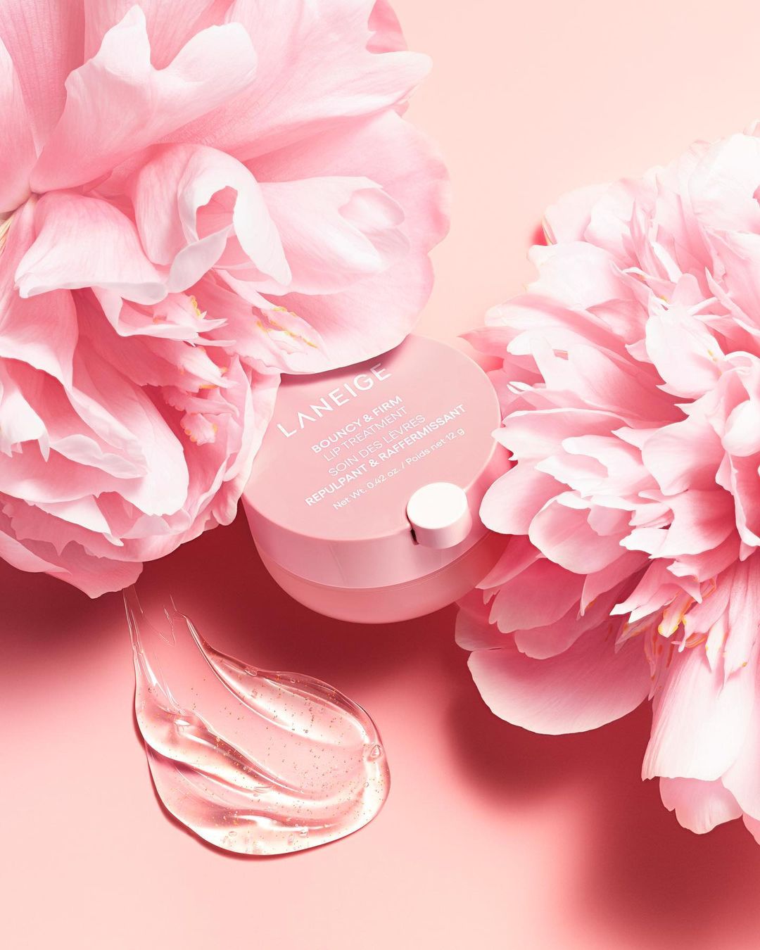Laneige skincare product sits amongst pink peony flowers with a smear of translucent gel on a pale pink surface in soft studio lighting.