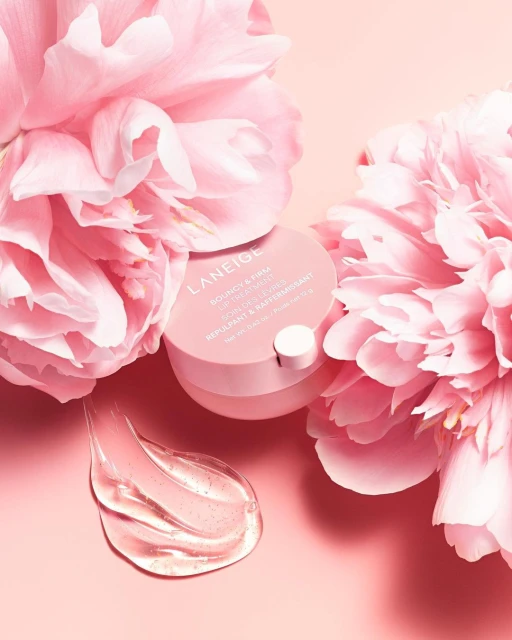 Laneige skincare product sits amongst pink peony flowers with a smear of translucent gel on a pale pink surface in soft studio lighting.