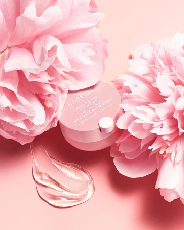 Laneige skincare product sits amongst pink peony flowers with a smear of translucent gel on a pale pink surface in soft studio lighting.