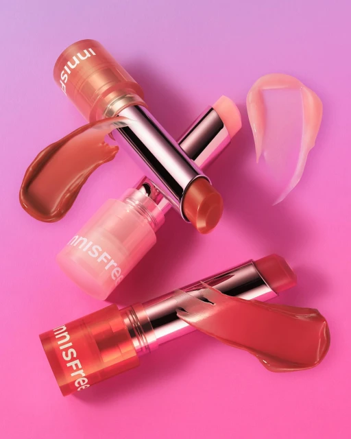 Close-up product shot of three Innisfree lip balms with colorful smears against a gradient pink and purple background. 