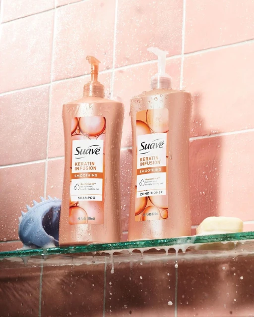 Two bottles of Suave Keratin Infusion shampoo and conditioner are displayed on a glass shelf against a pink tiled wall in a bathroom.