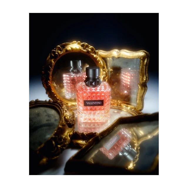 Valentino perfume bottle with geometric pattern and black cap sitting on a reflective surface with a gold ornate mirror background.