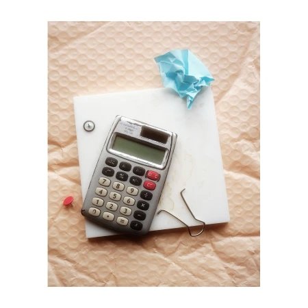 Overhead shot of calculator, paper clip, thumb tacks, and crumpled blue paper sitting on white board on top of bubble wrap.