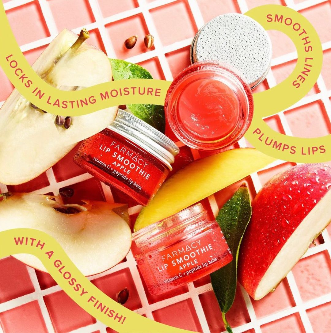 Close-up of Farmacy apple lip smoothie balm with sliced apple and pear on a pink tile surface, promoting smooth lines and lasting moisture.