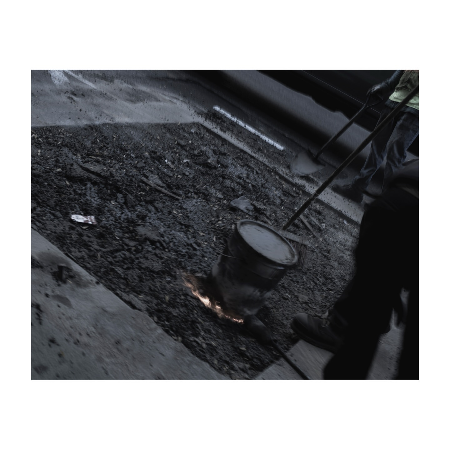 Construction worker performing road repair with molten material and a bucket in an urban environment, grayscale color palette.