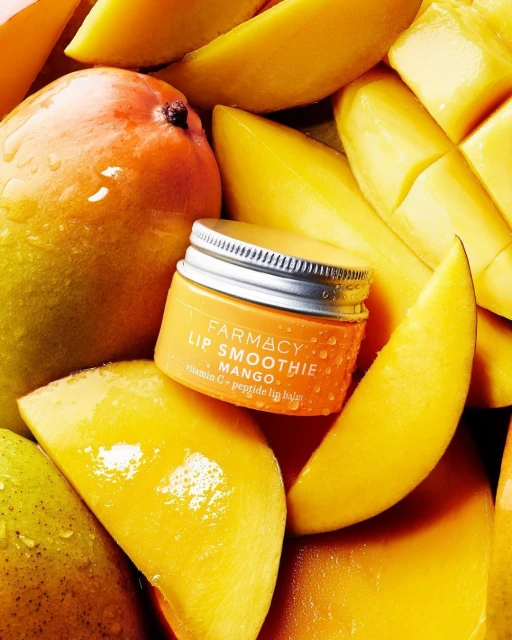 Close-up of Farmacy Lip Smoothie Mango lip balm surrounded by sliced mangos, showcasing beauty and skincare product with fresh fruit.
