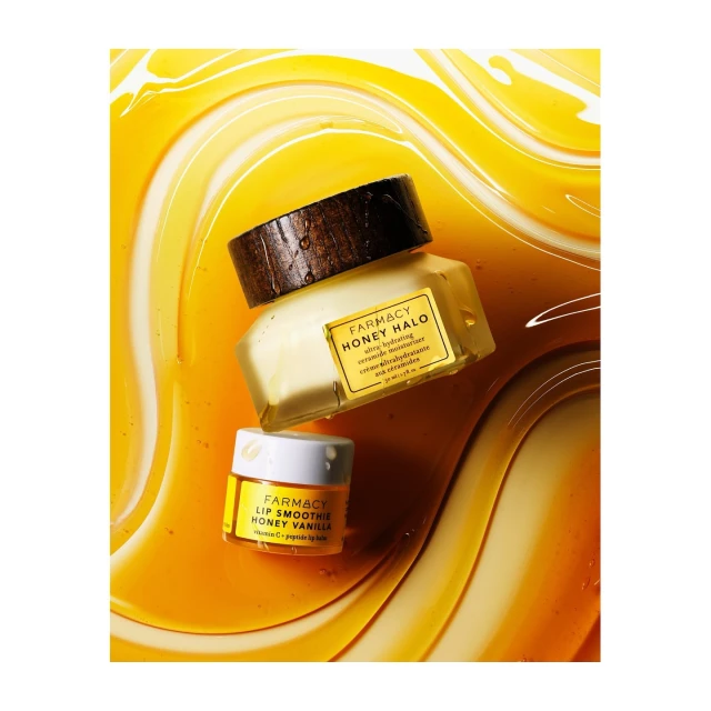 Two jars of Farmacy skincare products, Honey Halo and Lip Smoothie Honey Vanilla, surrounded by honey-like liquid on a white surface.