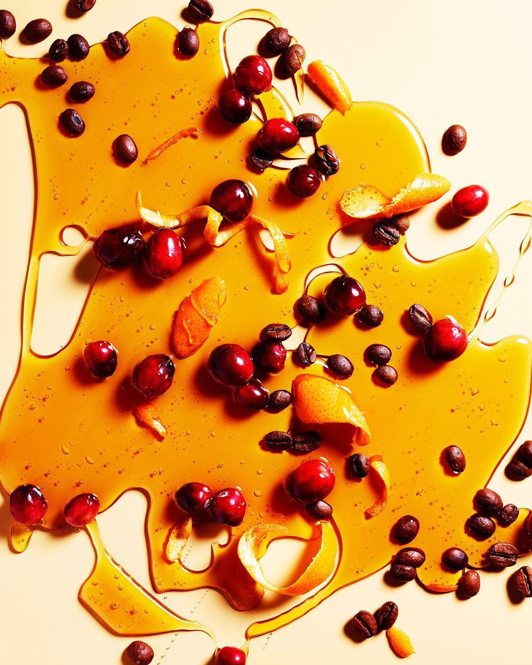 Overhead shot featuring a visually appealing arrangement of coffee beans, cranberries, orange peels, and a glossy honey-like liquid.