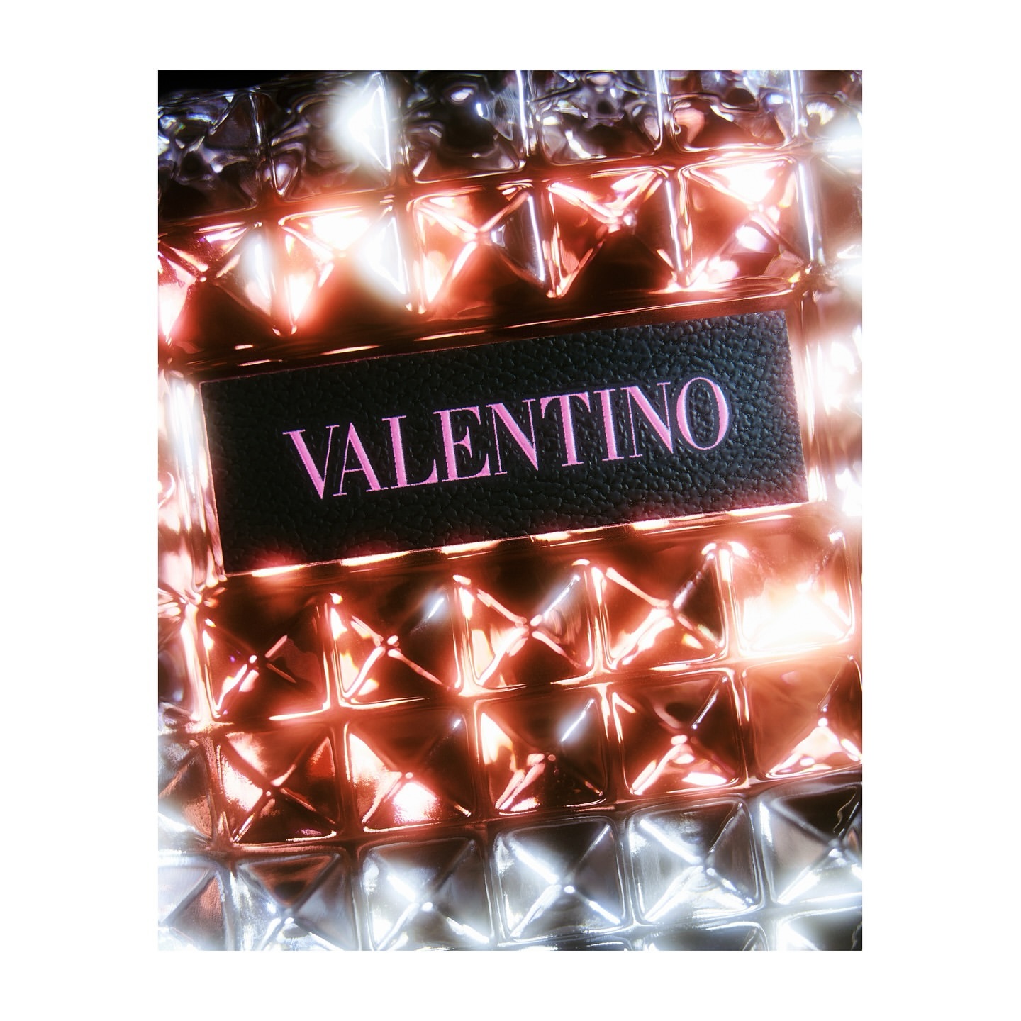Close-up studio shot of a Valentino textured rectangular perfume bottle with a black label, featuring bright pink text on a white background.