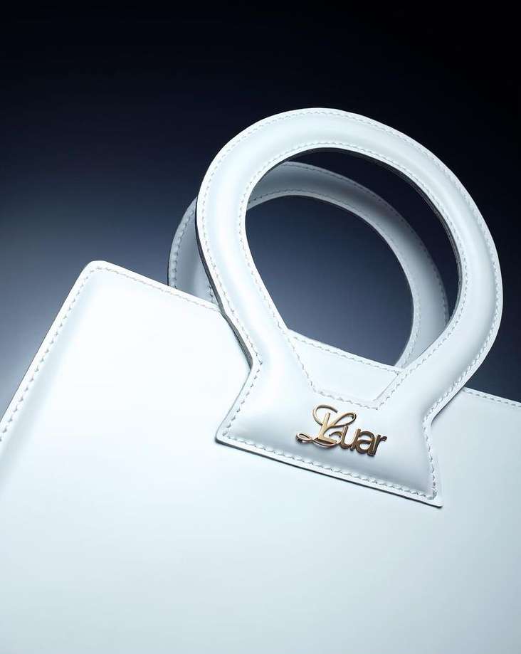 Close-up shot of a white leather handbag with a round handle and a gold 'Luar' logo against a dark background, studio lighting.