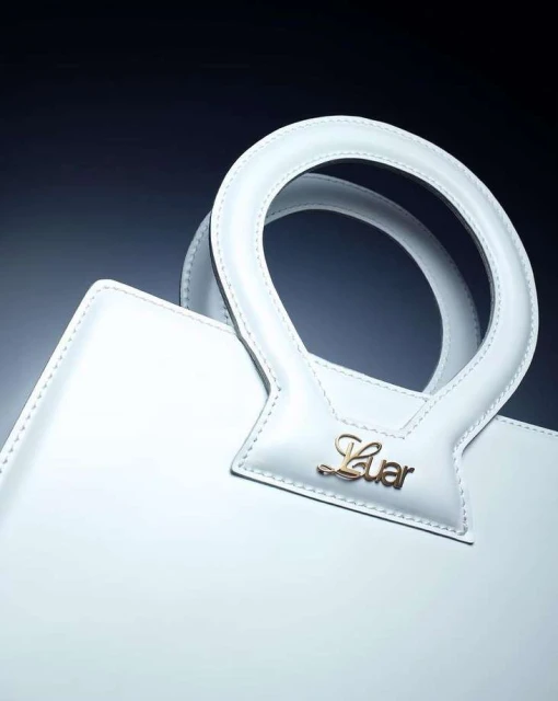 Close-up shot of a white leather handbag with a round handle and a gold 'Luar' logo against a dark background, studio lighting.