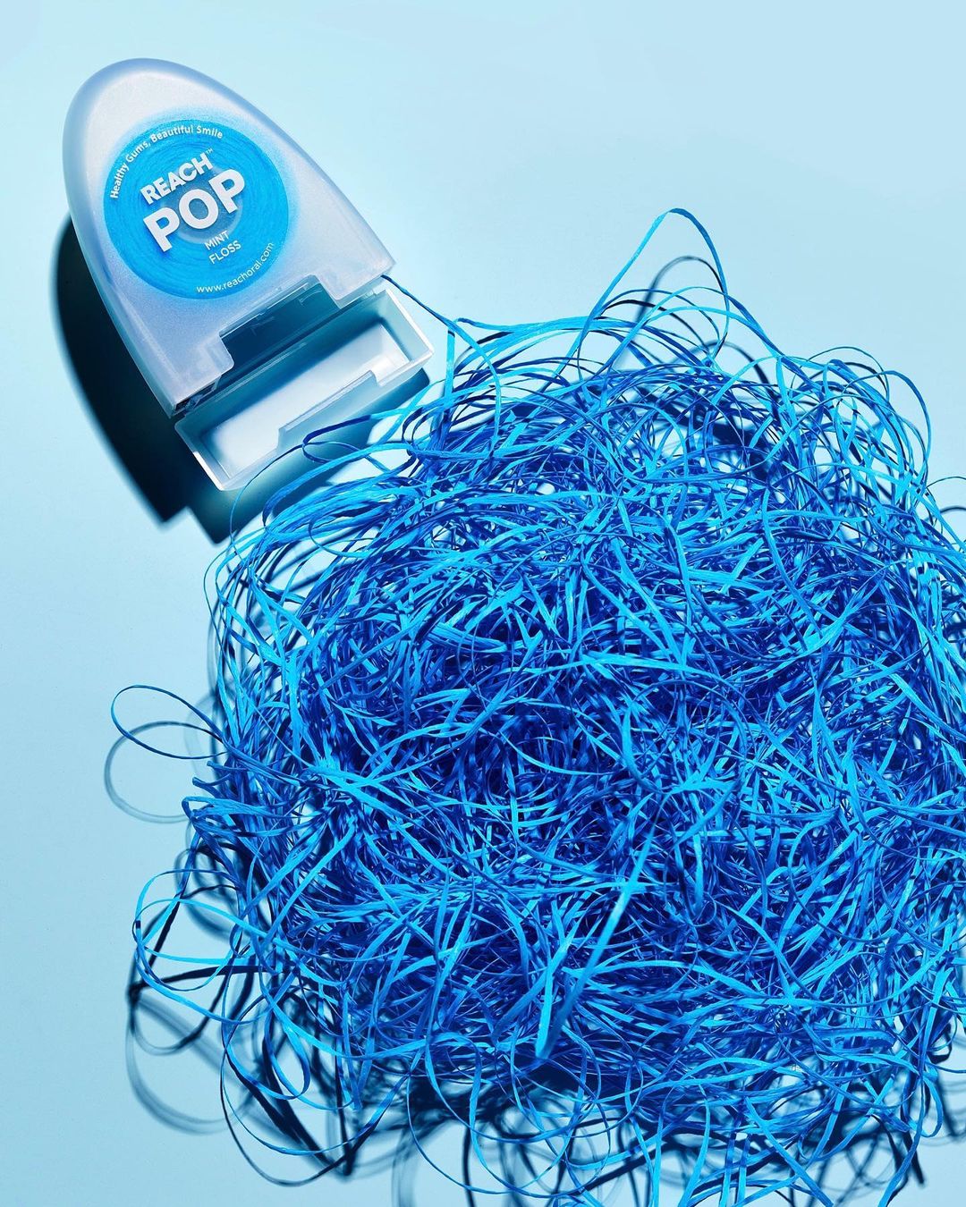 Reach Pop dental floss product with blue floss strands arranged on a light blue background in a brightly lit product shot.