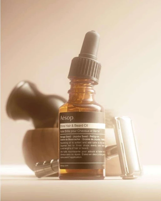 Close-up of amber glass bottle with Aesop Shine Hair & Beard Oil label, brush, and razor against a beige background in soft, diffused lighting.