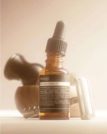 Close-up of amber glass bottle with Aesop Shine Hair & Beard Oil label, brush, and razor against a beige background in soft, diffused lighting.