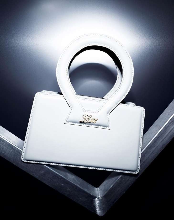 Close-up shot of a white leather handbag with a circular handle on a geometric metal surface against a blurred black background.