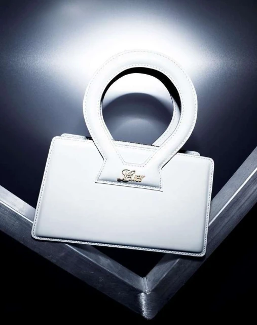 Close-up shot of a white leather handbag with a circular handle on a geometric metal surface against a blurred black background.