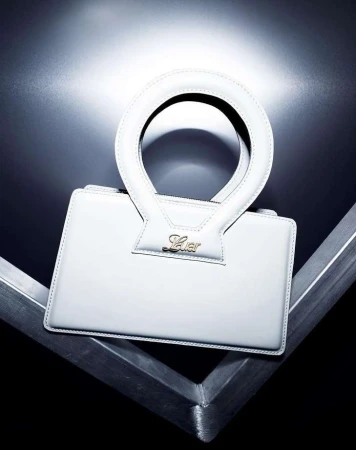 Close-up shot of a white leather handbag with a circular handle on a geometric metal surface against a blurred black background.