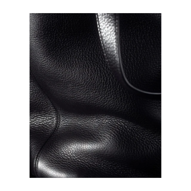 Close-up photograph of a black leather bag's surface showing its texture and a portion of its handle, set against a plain backdrop.