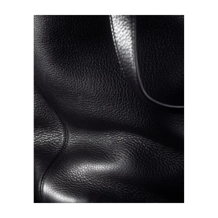 Close-up photograph of a black leather bag's surface showing its texture and a portion of its handle, set against a plain backdrop.