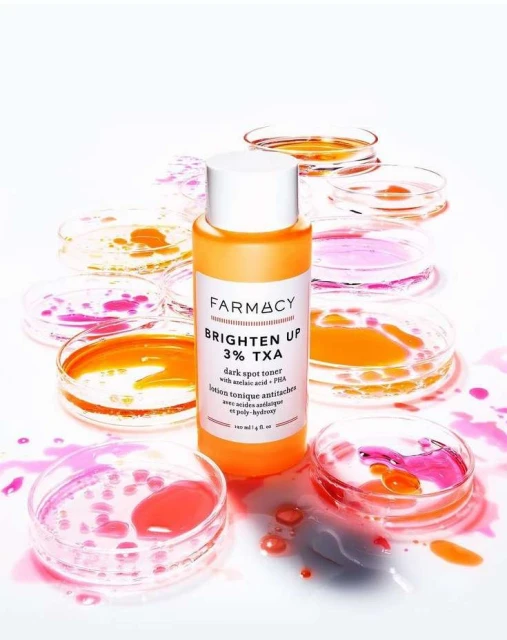 Farmacy Brighten Up 3% TXA dark spot toner bottle surrounded by petri dishes with colorful liquids on a clean white background.
