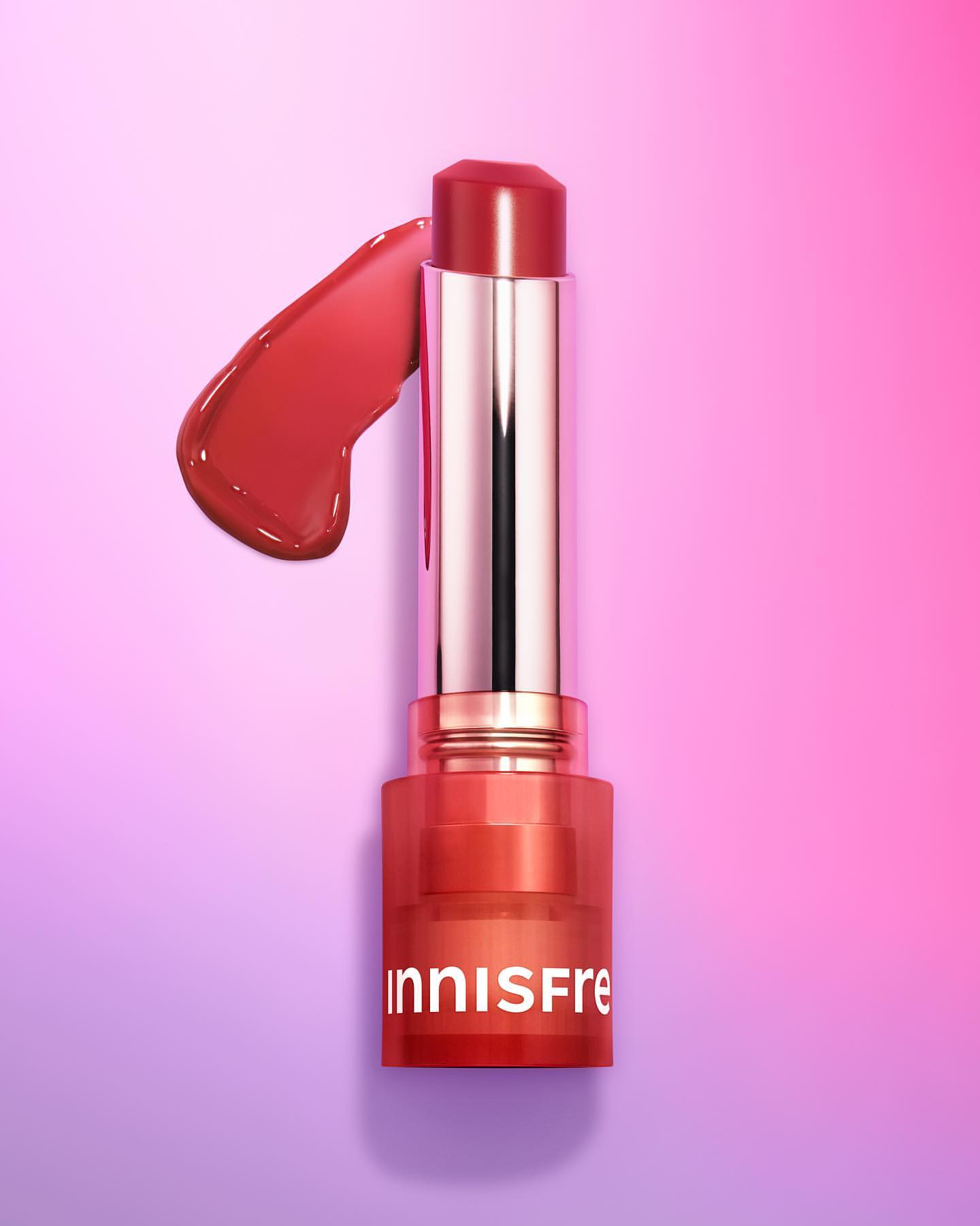 Close-up of Innisfree lip balm stick with a glossy swatch on a gradient pink and purple background. Makeup, cosmetic product shot.