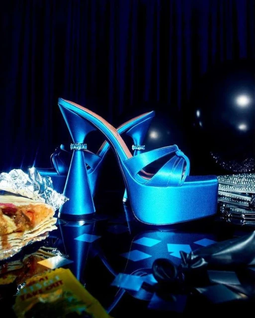 Turquoise platform high-heeled shoes with embellished ankle straps against a dark background with food props in a fashion editorial.
