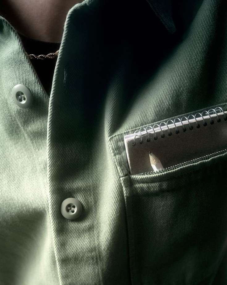 Close-up of a light green button-up shirt with a notebook and pencil sticking out of the pocket on the right side of the frame.