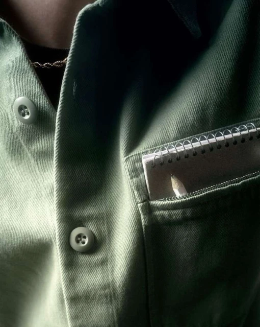 Close-up of a light green button-up shirt with a notebook and pencil sticking out of the pocket on the right side of the frame.