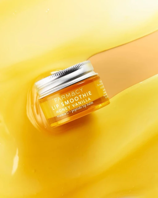 Close-up of Farmacy Lip Smoothie Honey Vanilla lip balm in a glass jar, set against a swirling yellow and cream background. Beauty product shot.