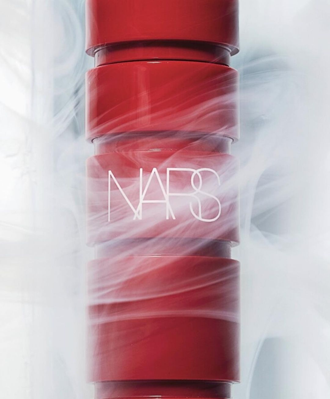 Close-up of a red NARS lipstick tube with the logo visible and swirling white smoke in the background against a white background.