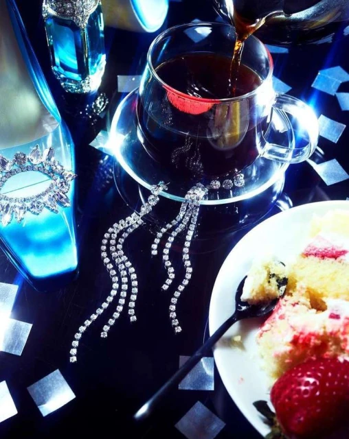 Pouring dark liquid into a glass cup on a table with a slice of cake, jewelry, and a blue high-heeled shoe.