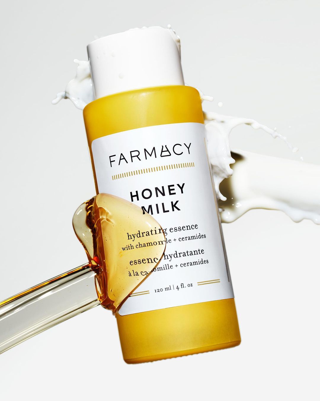 Farmacy Honey Milk hydrating essence bottle on white backdrop with milk splash and clear liquid on a clear stirring rod.
