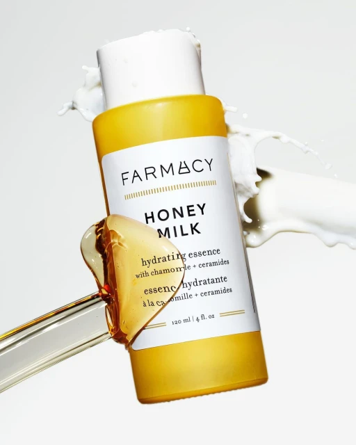 Farmacy Honey Milk hydrating essence bottle on white backdrop with milk splash and clear liquid on a clear stirring rod.