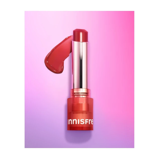 Close-up of Innisfree lip product with reflective packaging, accompanied by a smear of the lipstick on a pink and purple gradient background.