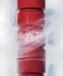 Close-up product shot of a red NARS cosmetic tube surrounded by wispy white smoke against a pale background