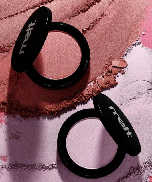 Two round black cosmetic compacts with open lids resting on swatched pink and purple powder makeup backgrounds.