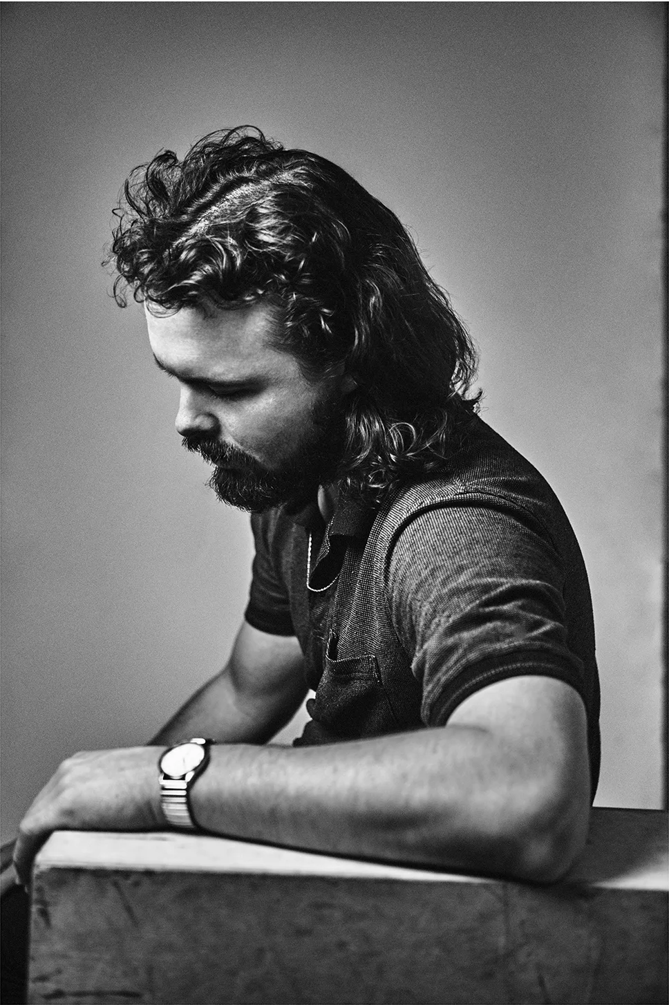 Black and white portrait of a man with a beard and mullet hairstyle, wearing a collared shirt, sitting with his arms resting on a box.