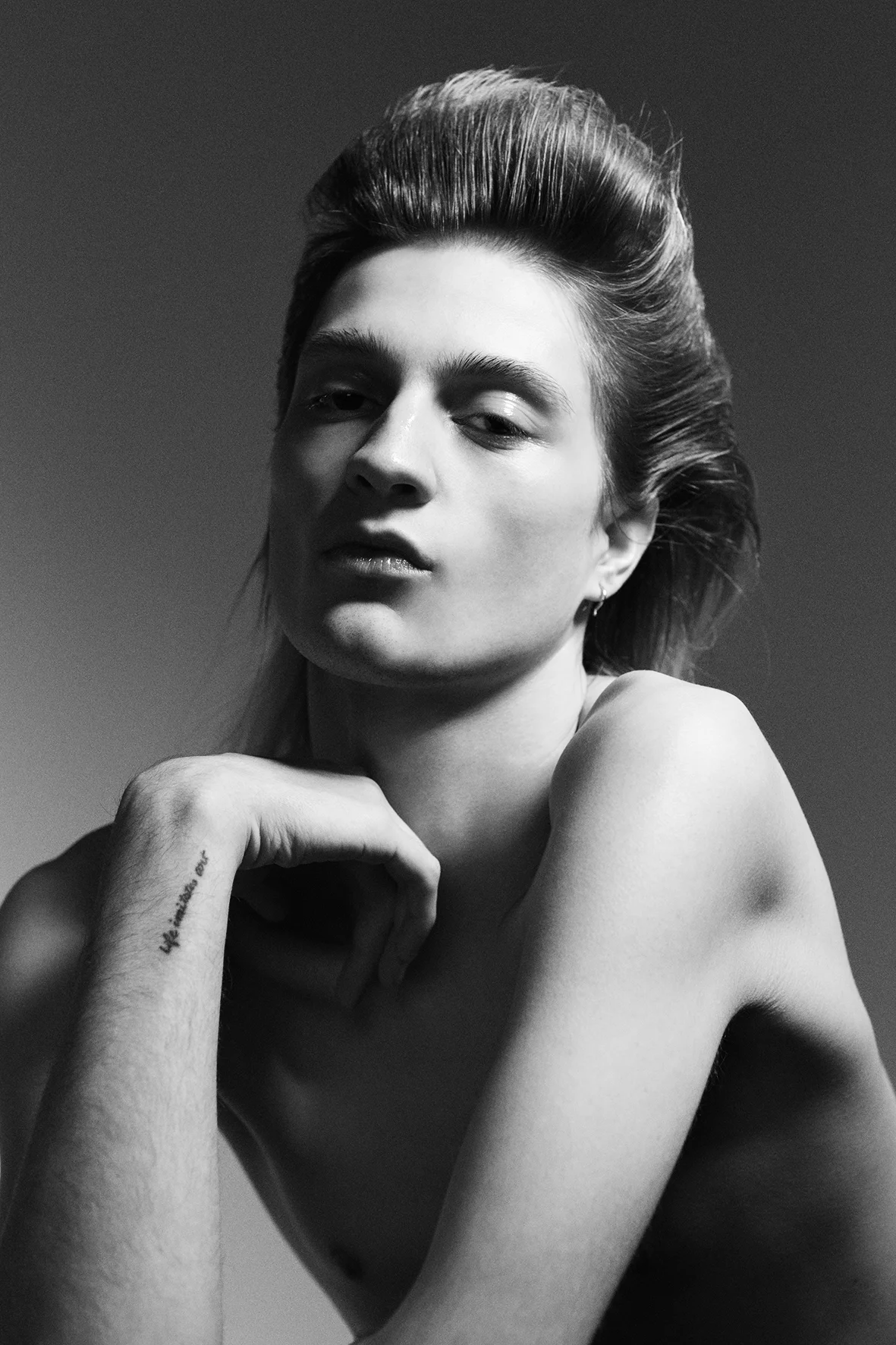 Black and white portrait of a young adult with styled hair, visible tattoo on arm, set against a neutral gradient background.