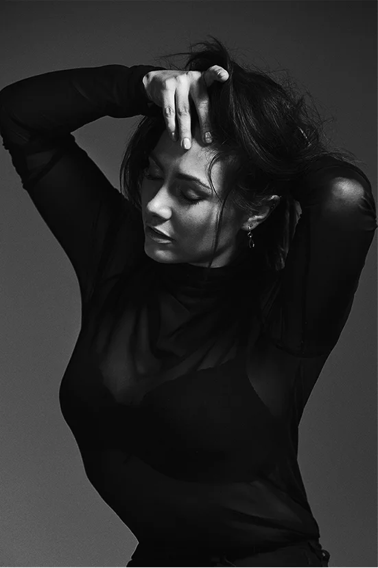Black-and-white portrait of a woman with dark hair in a black top, posing with her hands behind her head against a grey backdrop.