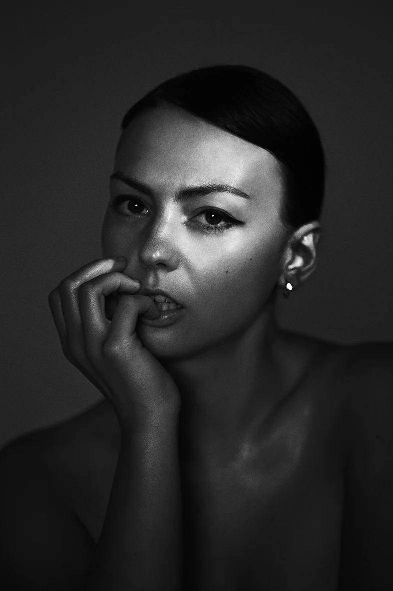Portrait of a woman in black and white with dark hair slicked back, hand near face and fingers near her mouth, looking at camera.