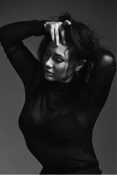 Monochrome portrait of a woman in a sheer black top running her hands through her dark hair against a plain grey background.