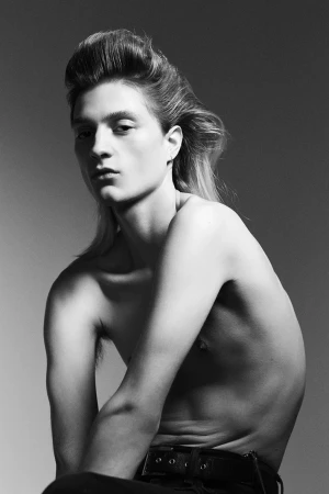 Monochrome portrait of a young male model with swept-back hair and a dark belt against a grey gradient studio background.