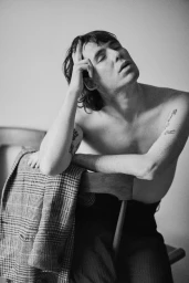 Black and white portrait of a shirtless man leaning on a wooden chair with a plaid jacket draped over the back.