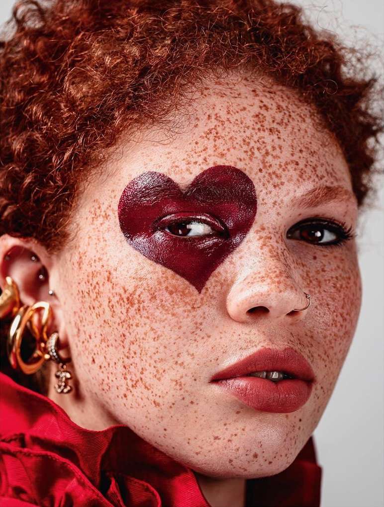 Close-up portrait of a red-haired woman with a heart painted over her eye, wearing a red top and gold earrings against a grey backdrop.