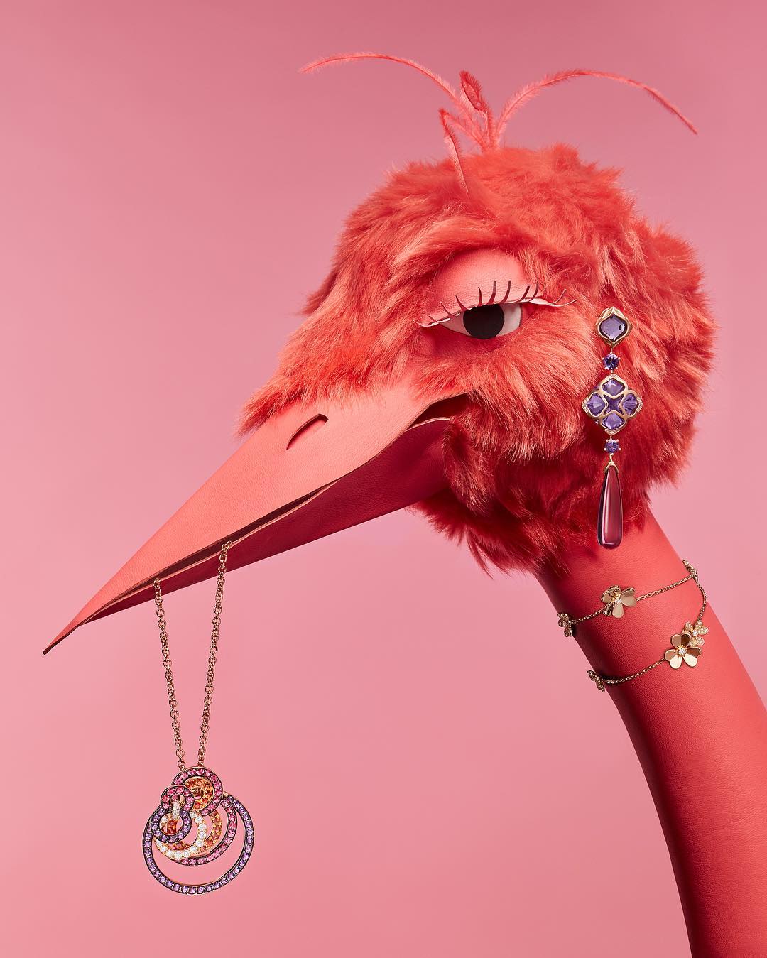 Whimsical fashion shot of a coral-pink bird puppet adorned with jewelry, including earrings, necklace, and bracelet, against a pink backdrop.