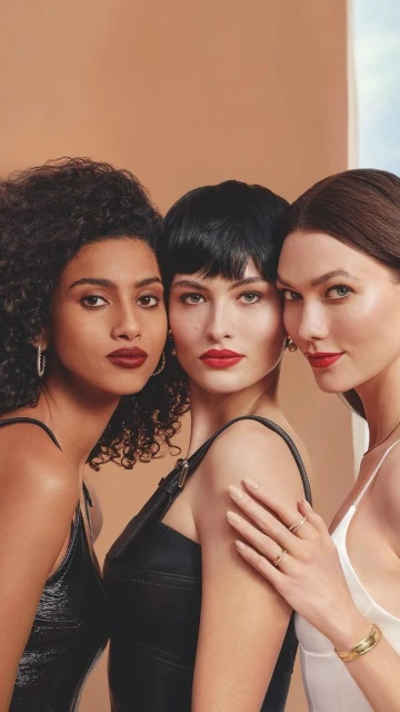 Close-up of three adult women with dark red lipstick, jewelry, and neutral dresses in an indoor setting with a tan and blue background.