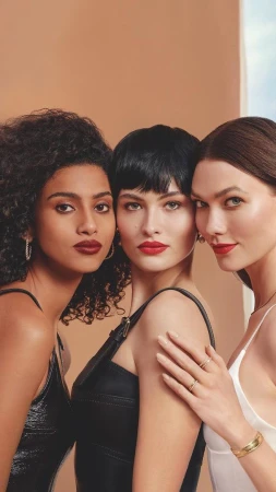 Close-up of three adult women with dark red lipstick, jewelry, and neutral dresses in an indoor setting with a tan and blue background.