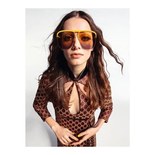 A woman wearing oversized yellow sunglasses and a patterned dress poses against a bright white backdrop in a fashion editorial.