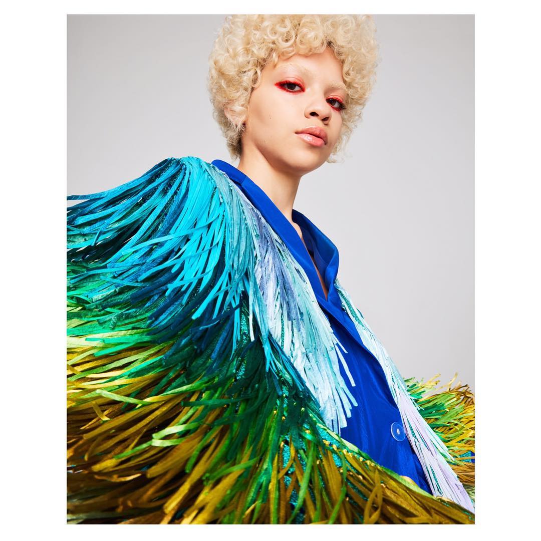 Woman with blonde curly hair and red makeup wears a blue silk shirt and a fringe jacket with blue, green, and yellow colors.