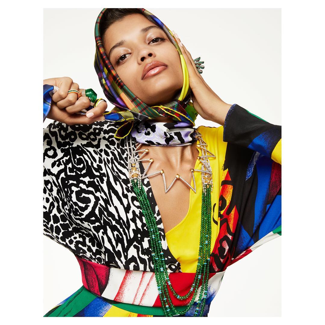 A stylish woman with a patterned headscarf and a colorful geometric-patterned dress poses indoors with jewelry and rings.